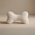 A cream bone-shaped pet toy with a small Woolmark black tag, resting on a neutral beige surface against a matching beige background.