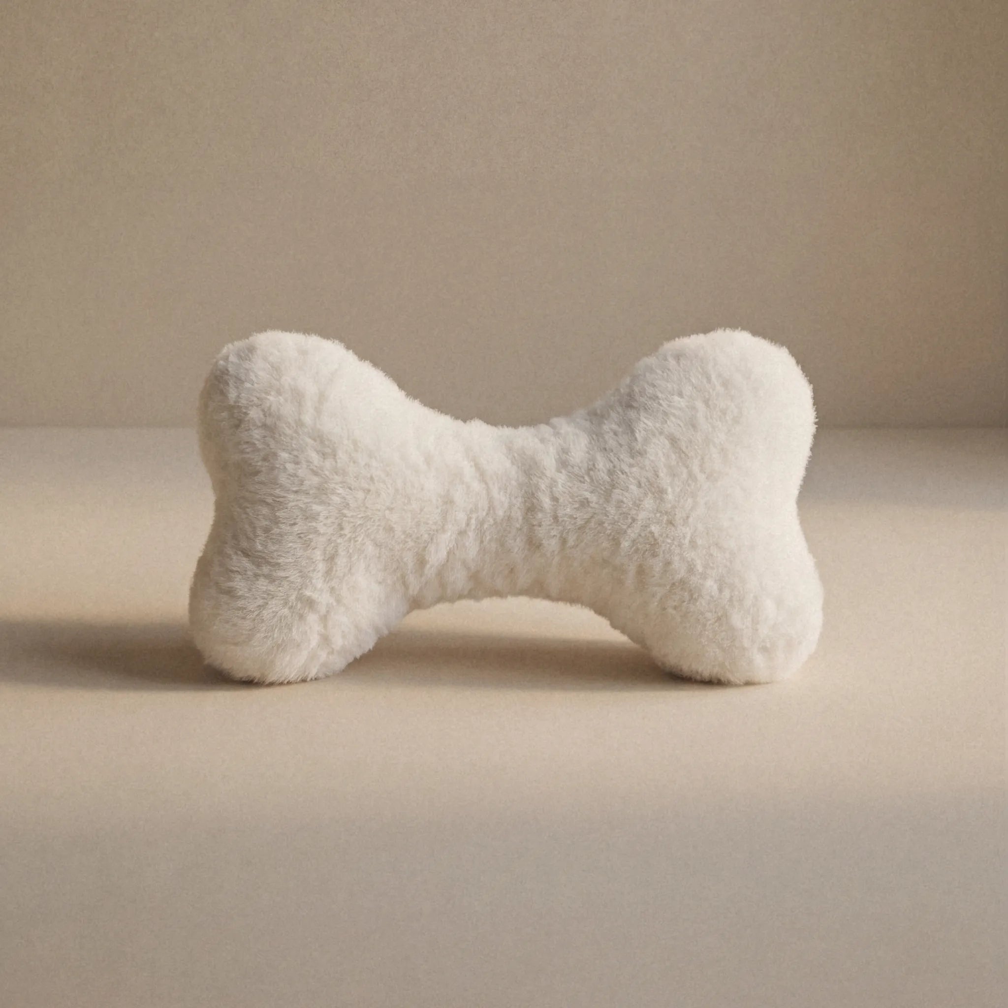 A cream bone-shaped pet toy with a small Woolmark black tag, resting on a neutral beige surface against a matching beige background.