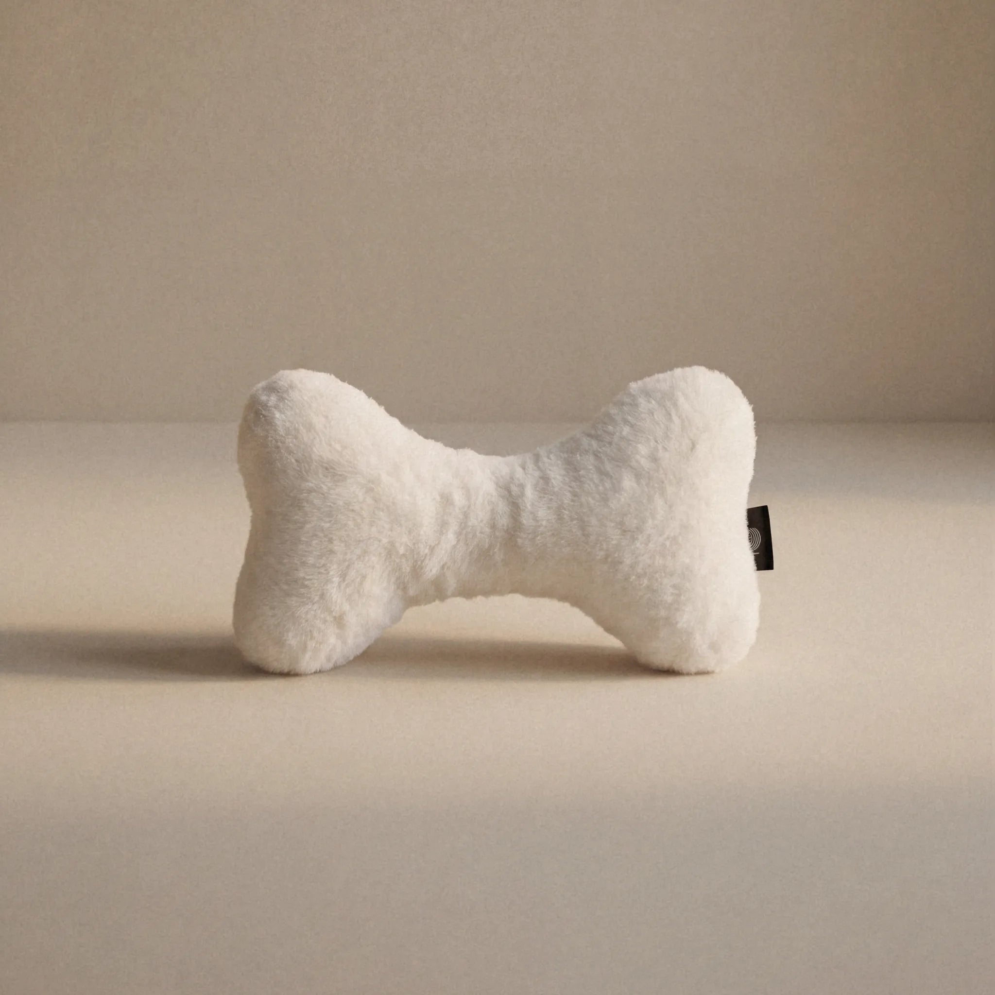 A cream bone-shaped pet toy with a small Woolmark black tag, resting on a neutral beige surface against a matching beige background.