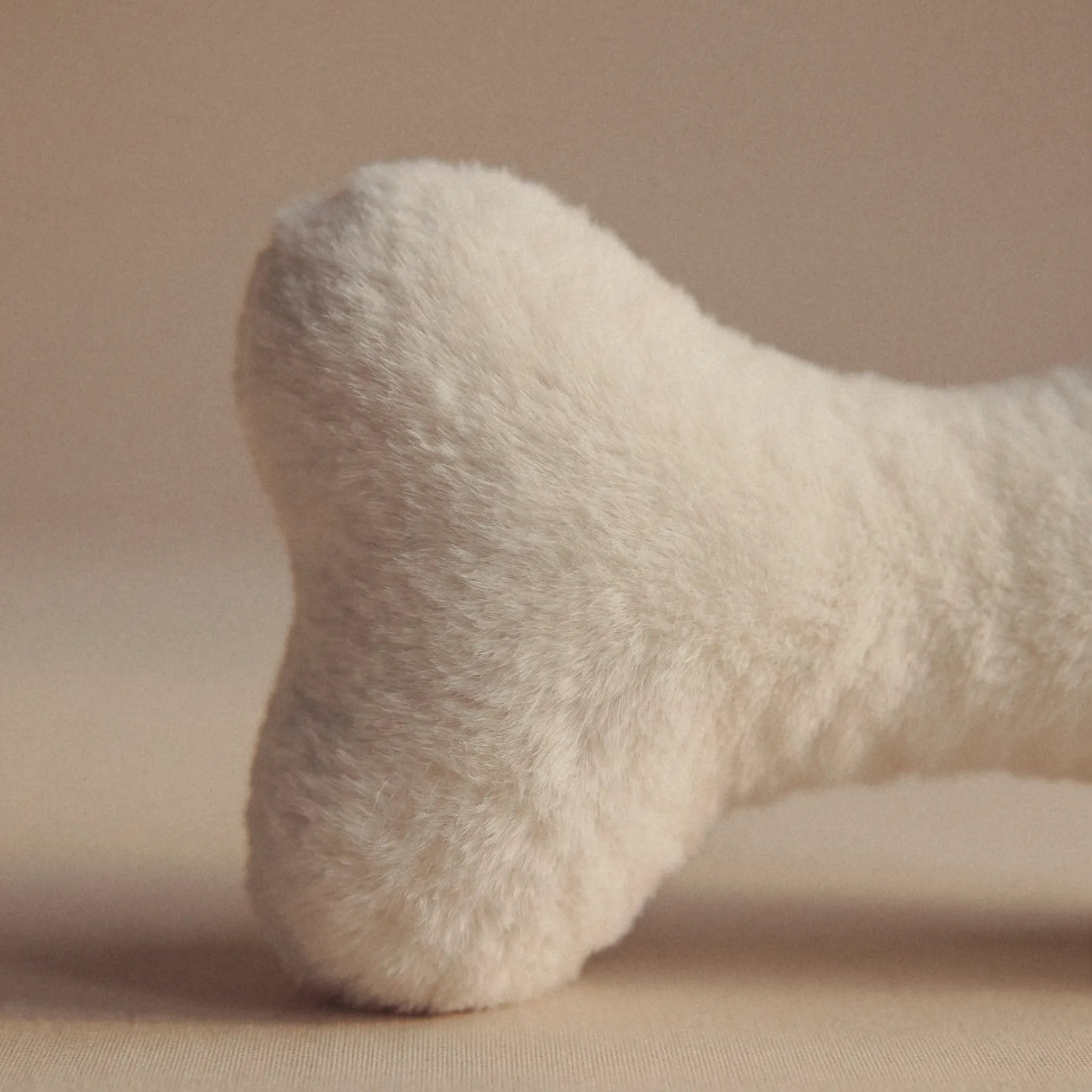 A close-up of a soft, bone-shaped plush pet toy in a warm off-white or cream color, set against a solid beige background.