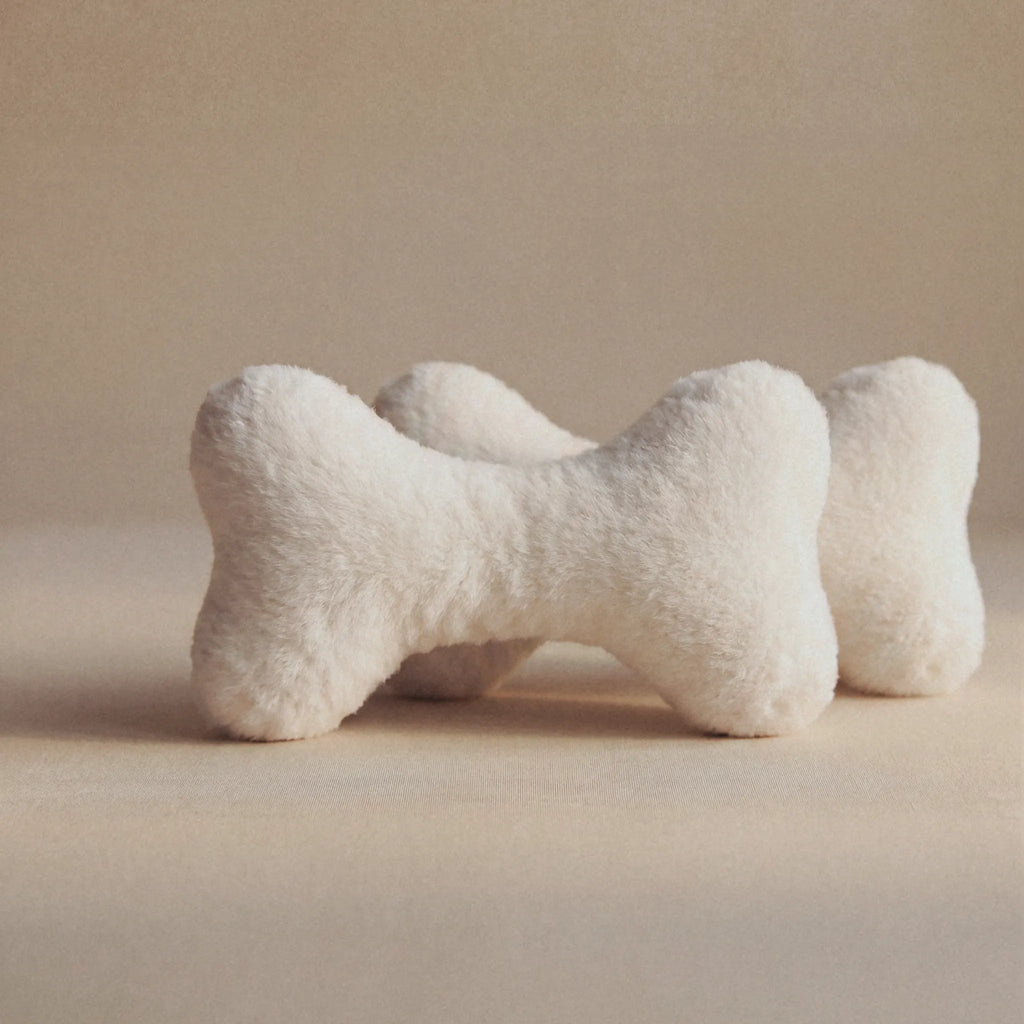 Two plush bone-shaped pet toys in a soft off-white faux fur material, arranged on a neutral beige background.