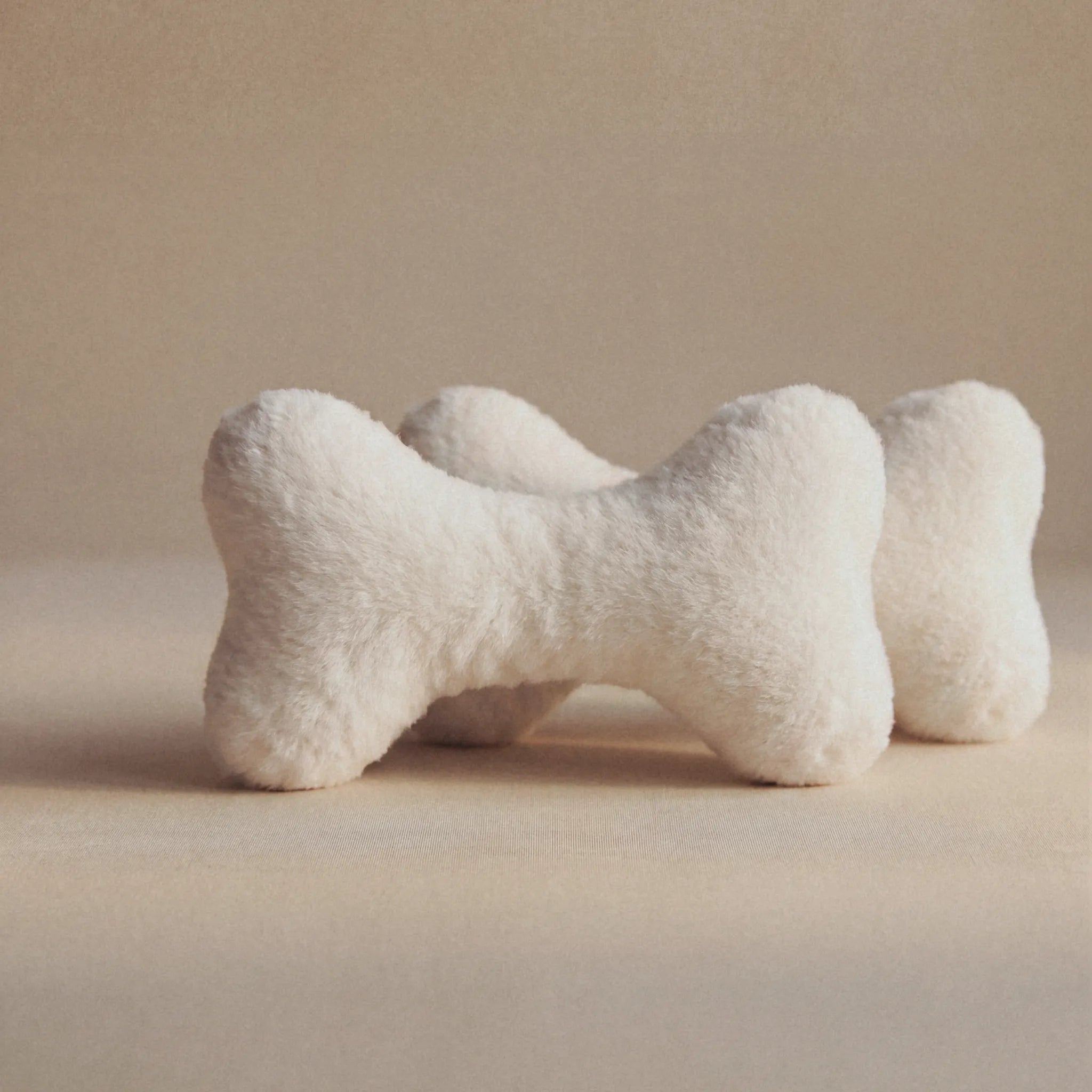Two plush bone-shaped pet toys in a soft off-white faux fur material, arranged on a neutral beige background.