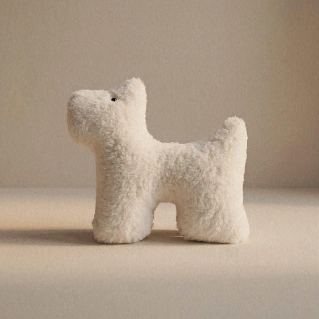 A small cream dog shaped plush pet toy made of cashmere standing on a plain beige surface against a matching beige background.