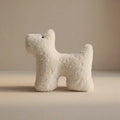 A small cream dog shaped plush pet toy made of cashmere standing on a plain beige surface against a matching beige background.