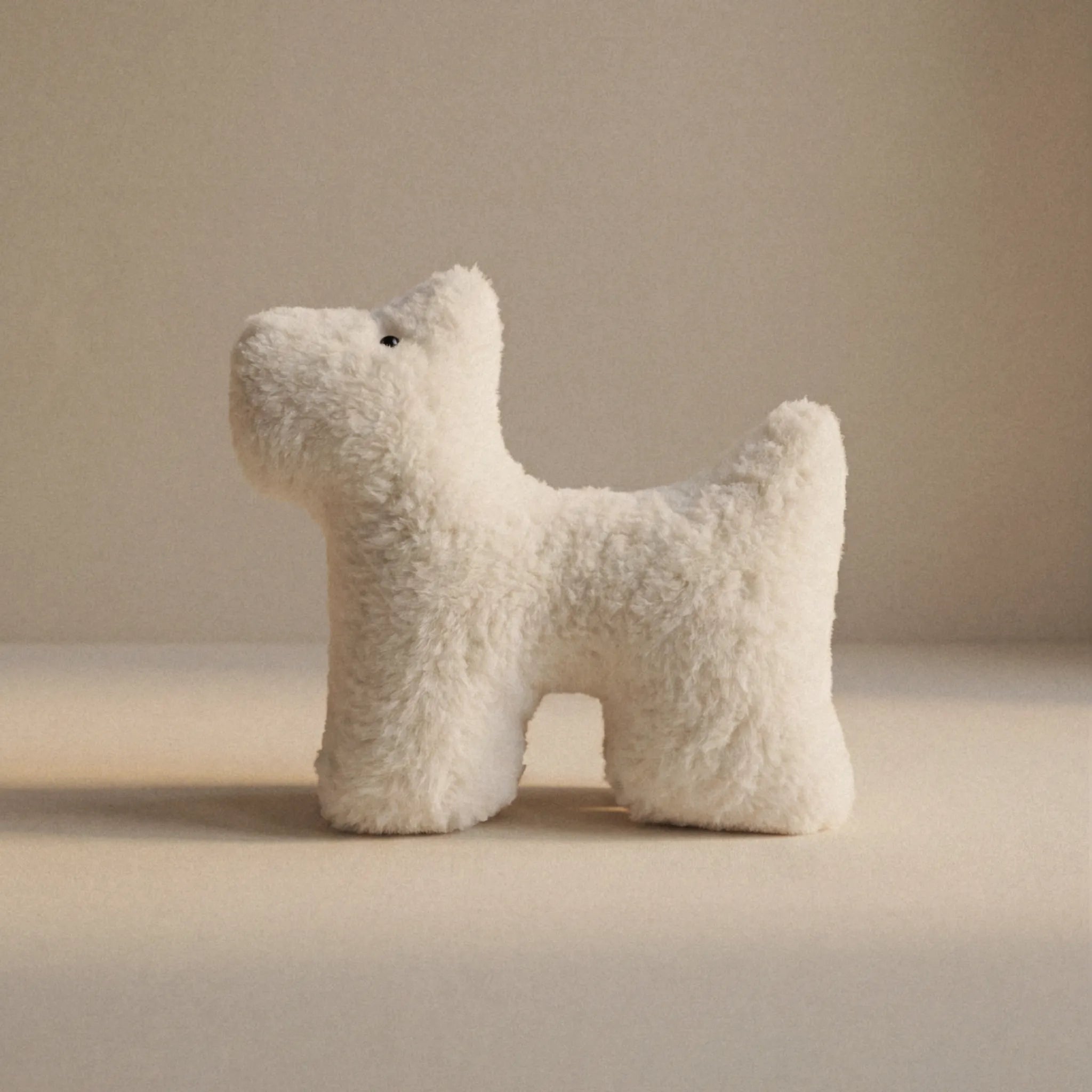 A small cream dog shaped plush pet toy made of cashmere standing on a plain beige surface against a matching beige background.