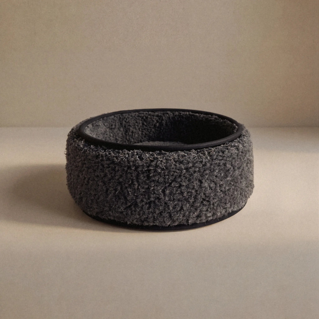 A dark wool sheep bouclé pet bed, with a black interior and base, photographed on a neutral beige surface and background.