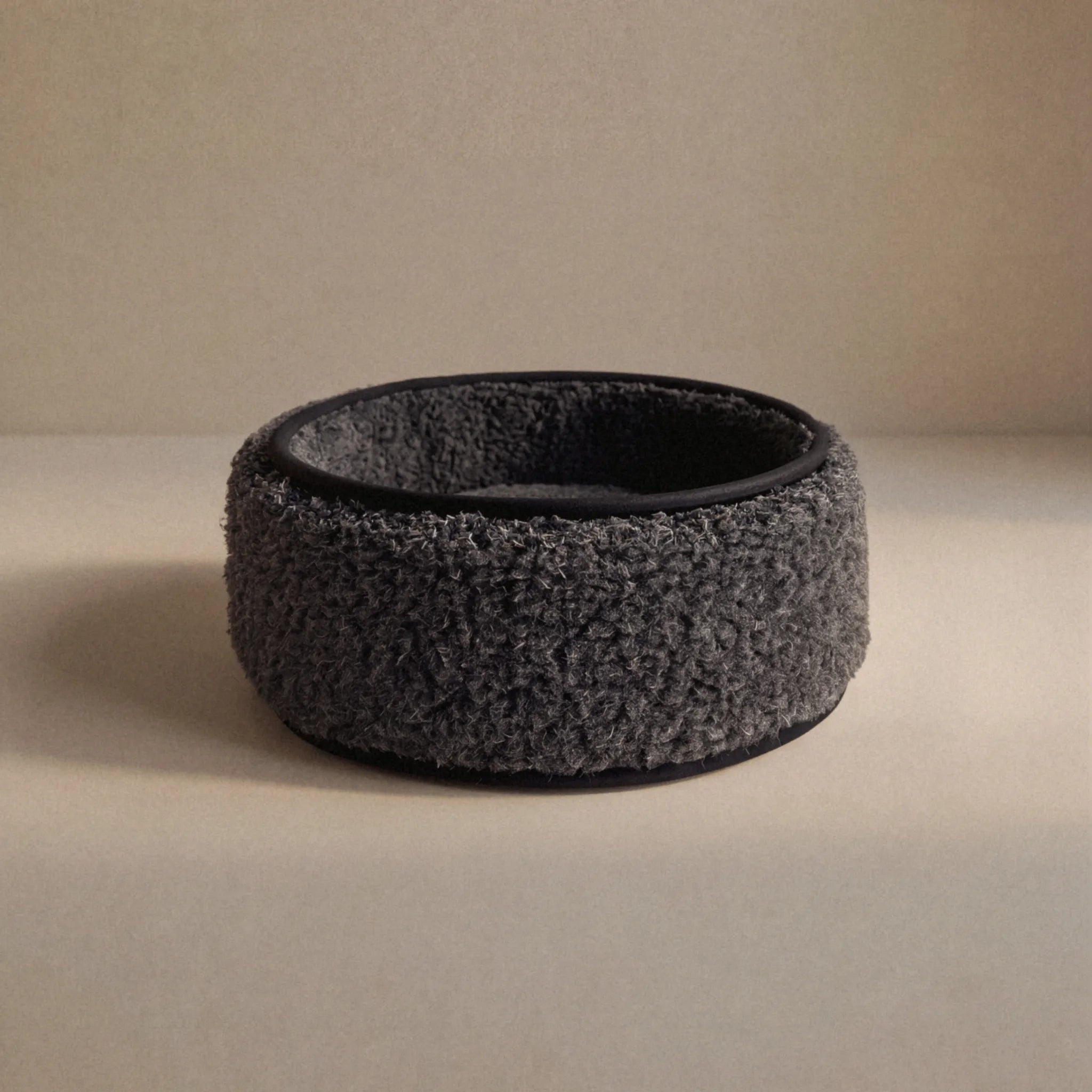 A dark wool sheep bouclé pet bed, with a black interior and base, photographed on a neutral beige surface and background.
