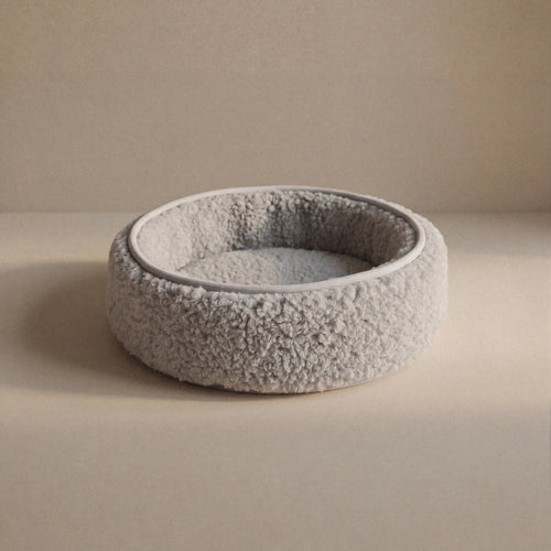 A round, wool sheep bouclé pet bed, with a textured, wool exterior and a soft, cushioned interior, shown on a neutral beige background.