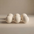 Six off-white wool balls arranged in two rows on a neutral beige surface against a matching beige background.