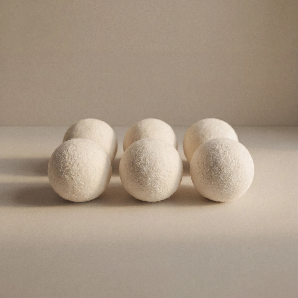 Six off-white wool balls arranged in two rows on a neutral beige surface against a matching beige background.