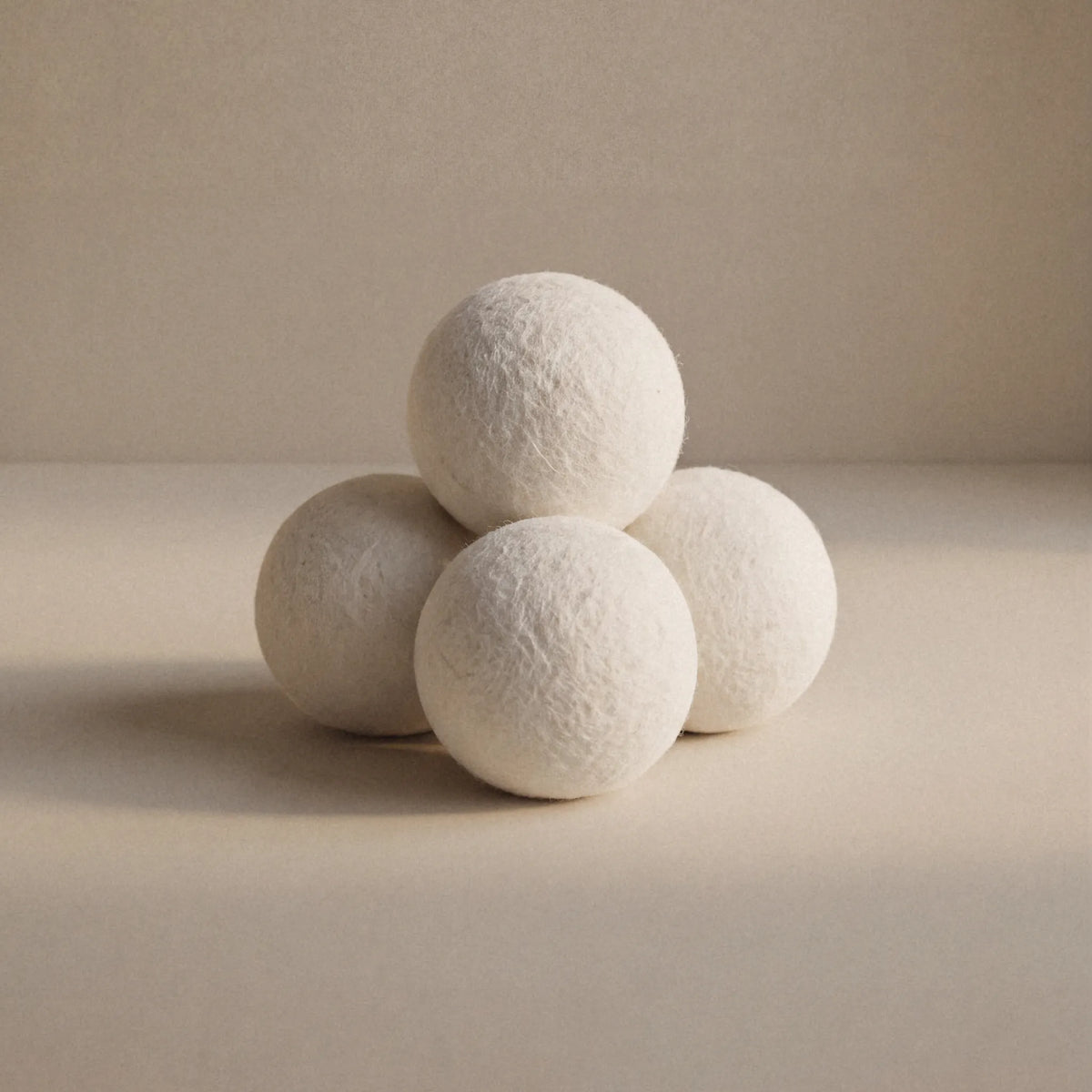 Four textured white wool balls arranged in a pyramid on a warm beige surface against a matching beige background.