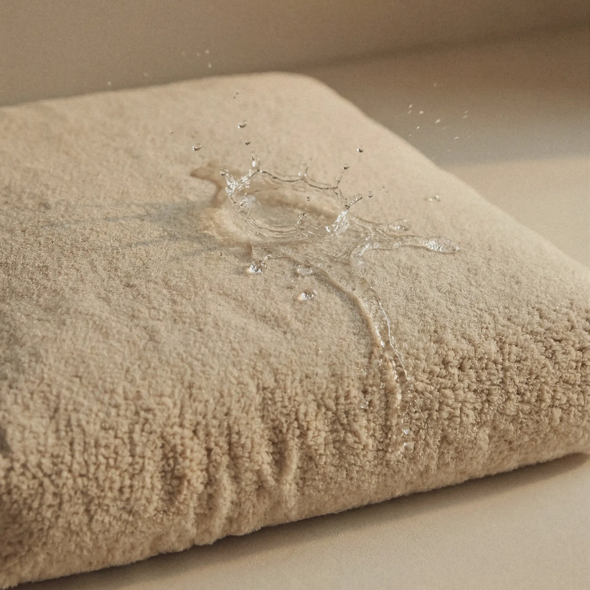 A close-up of a beige pet bed with water droplets and a small water splash on its surface, demonstrating a water-resistant or waterproof finish.